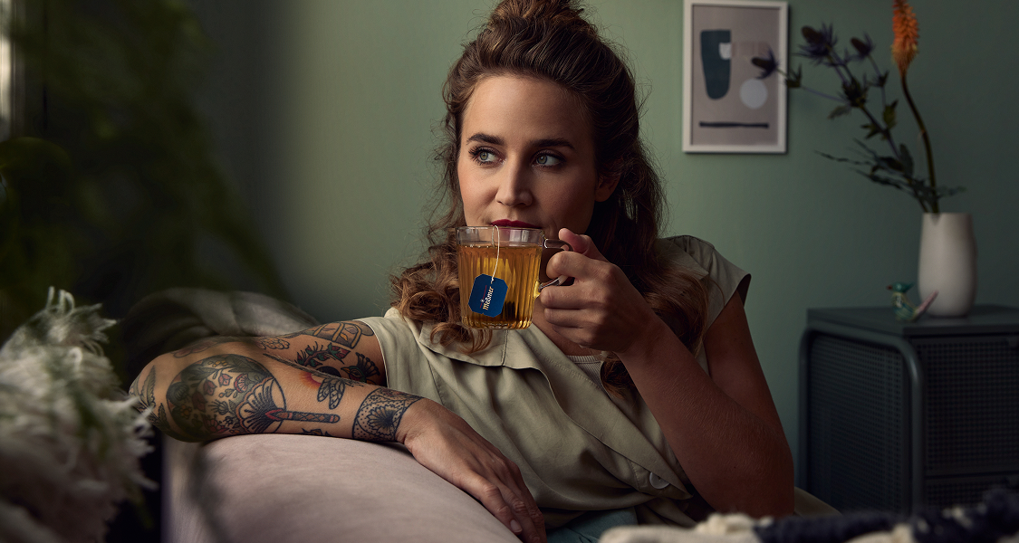 Eine Frau mit Tätowierungen sitzt auf einem Sofa und trinkt Tee aus einem Glasbecher. Sie schaut nachdenklich zur Seite in einem gemütlichen, sanft beleuchteten Raum mit gerahmter Kunst und Pflanzen.