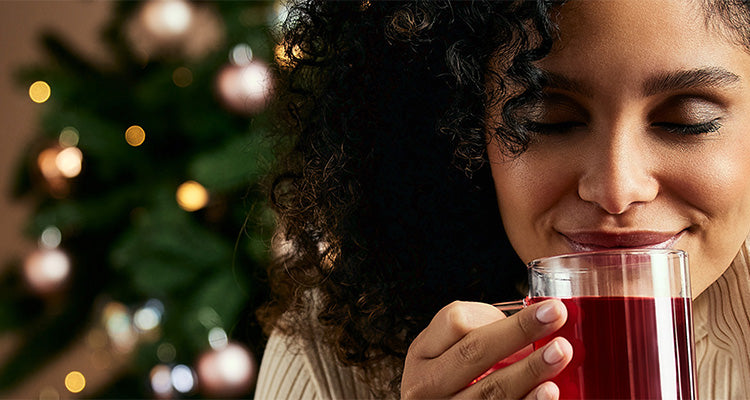 Eine Frau mit lockigem Haar lächelt mit geschlossenen Augen, während sie eine Glastasse mit rotem Tee in der Hand hält. Im Hintergrund ist ein geschmückter Weihnachtsbaum zu sehen.