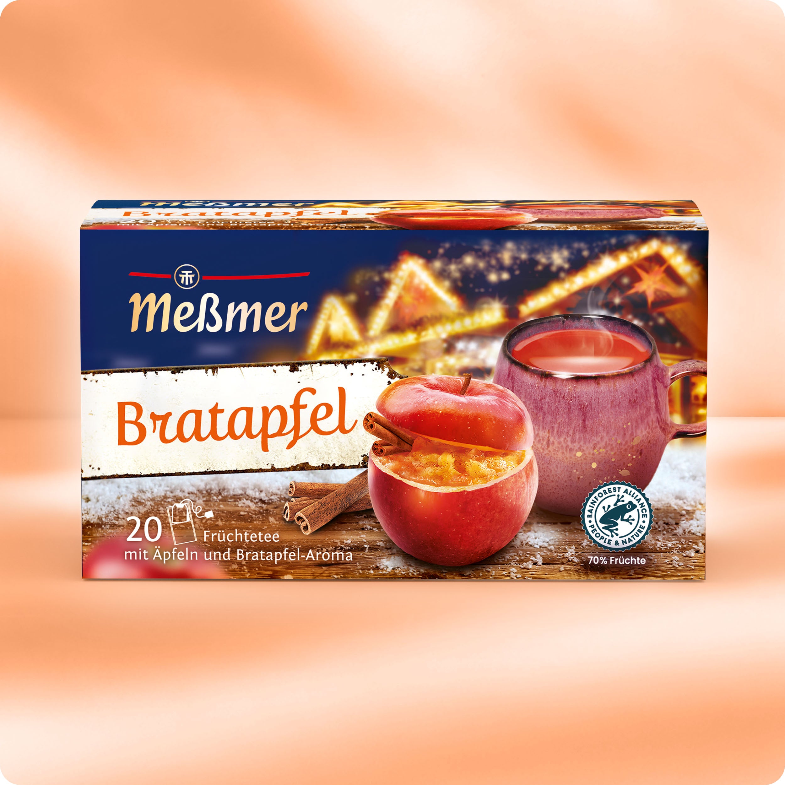 Eine Schachtel Meßmer DE Bratapfel Früchtetee, inspiriert vom Weihnachtsmarkt, zeigt eine warme, orangefarbene, festliche Verpackung mit verschwommenen, glitzernden Lichtern und einer Glastasse mit Tee neben einem Bratapfel.