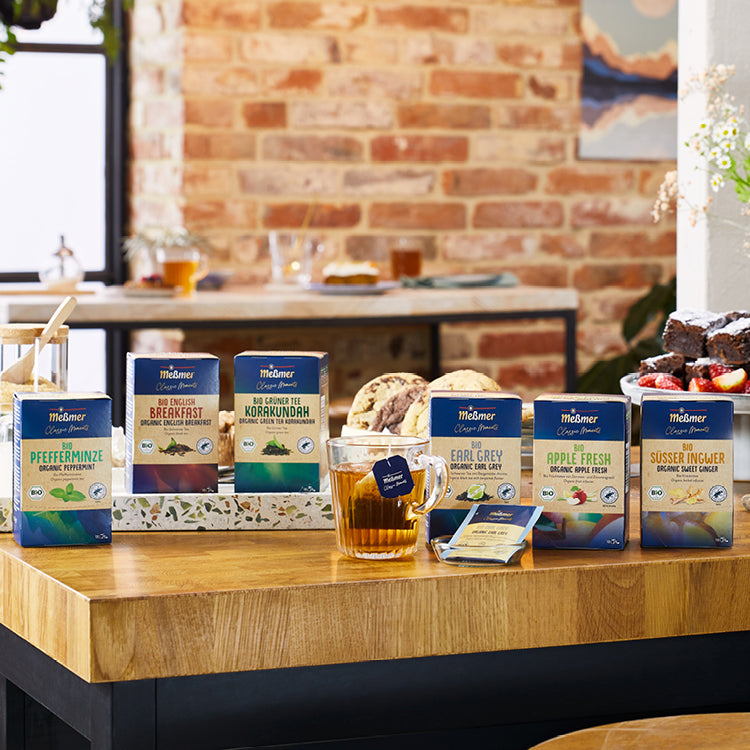Sechs Meßmer Teepackungen wie Pfefferminze, Breakfast, Korkundah, Earl Grey, Apple Fresh und Süßer Ingwer auf Holztablett, davor Glas mit Tee und Keksen, Backsteingwand im Hintergrund.
