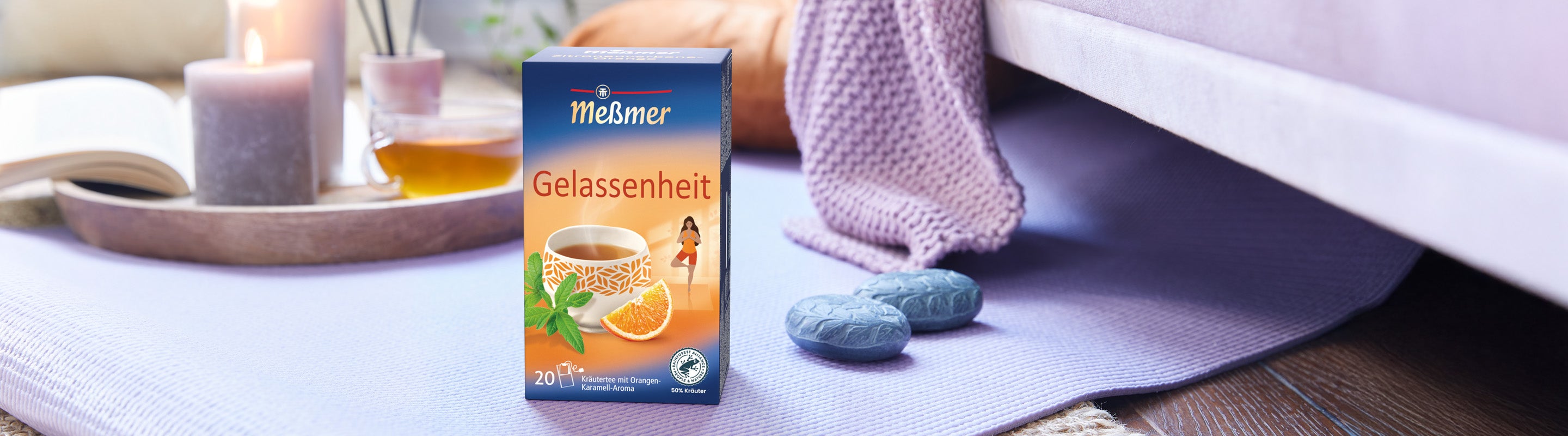Eine Schachtel Meßmer Gelassenheitstee liegt auf einer lila Matte neben blauen Steindekorationen, einer Decke und brennenden Kerzen und schafft eine ruhige, gemütliche Atmosphäre. Im Hintergrund ist auch ein aufgeschlagenes Buch zu sehen.