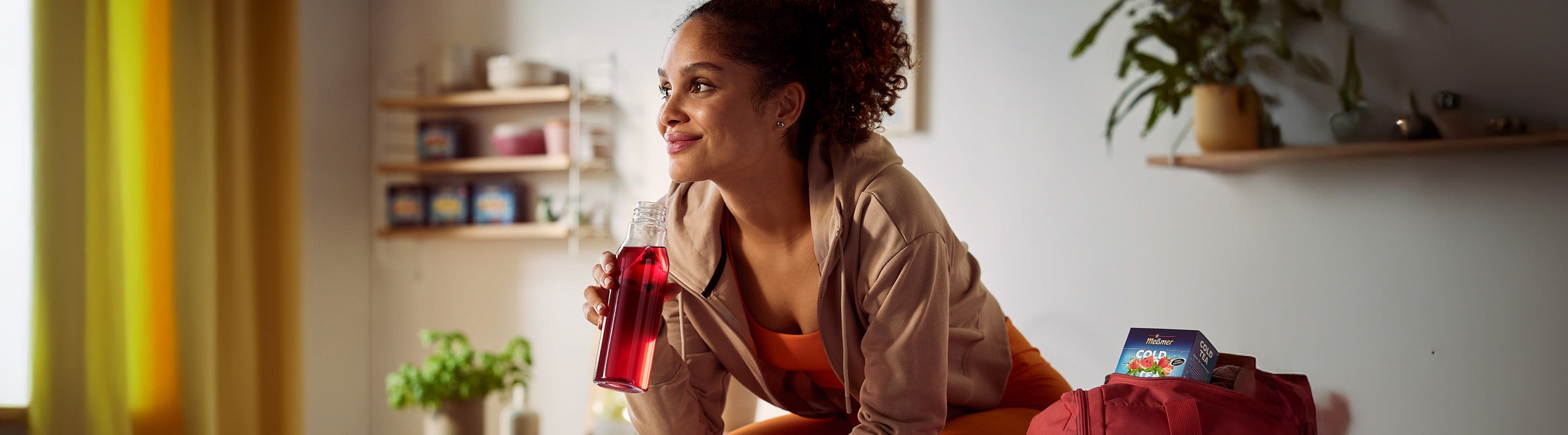 Eine Frau in Sportkleidung sitzt im Haus, lächelt und trinkt ein rotes Getränk aus einer durchsichtigen Flasche. Eine Sporttasche und eine Packung Saft stehen neben ihr. Der Raum ist hell und gemütlich, mit Regalen, Pflanzen und gelben Vorhängen im Hintergrund.