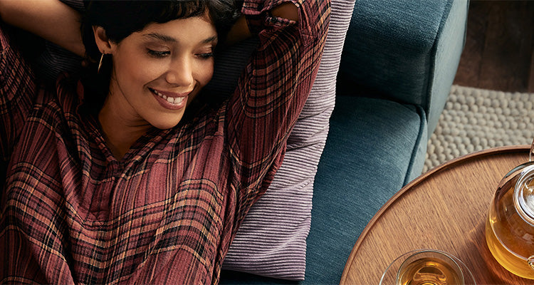 Eine Frau in einem rot karierten Hemd lehnt sich lächelnd und entspannt auf einem blauen Sofa mit einem Kissen zurück. Neben ihr steht ein Holztisch mit einer Teekanne und einer Tasse Tee.