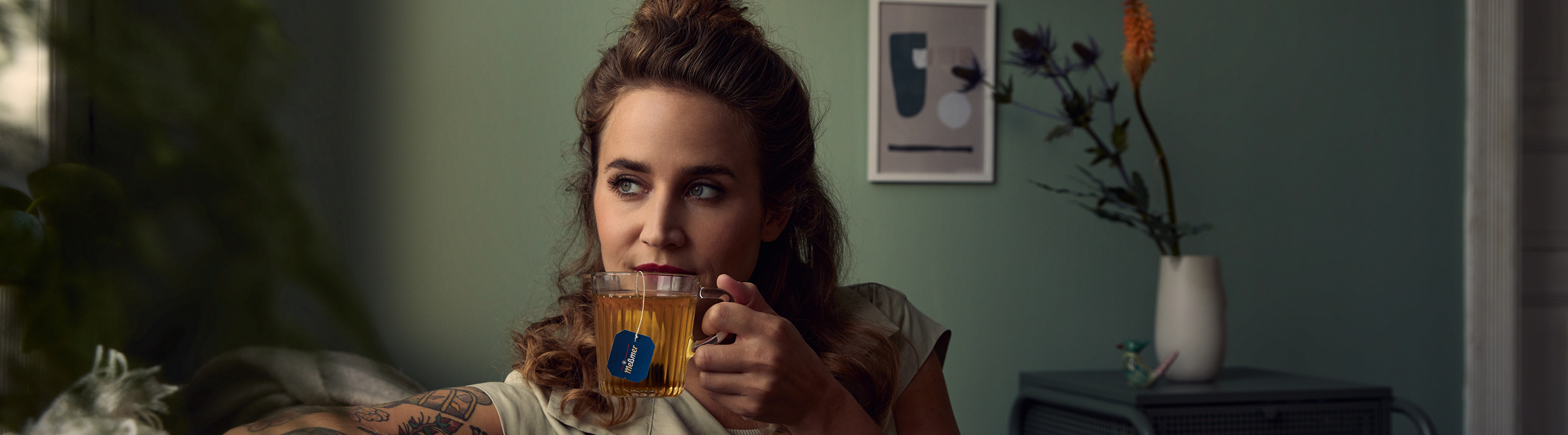 Eine Frau mit gewelltem braunem Haar sitzt in einem Haus und hält eine Glastasse mit einem blauen Teebeutel in der Hand. Sie blickt nachdenklich zur Seite. Eine Vase mit Blumen und ein gerahmtes Kunstwerk sind im sanft beleuchteten Hintergrund zu sehen.