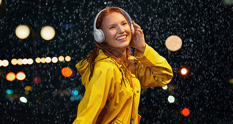 Eine lächelnde Frau in einem gelben Regenmantel hört Kopfhörer, während sie nachts im Regen steht, mit verschwommenen Stadtlichtern im Hintergrund.
