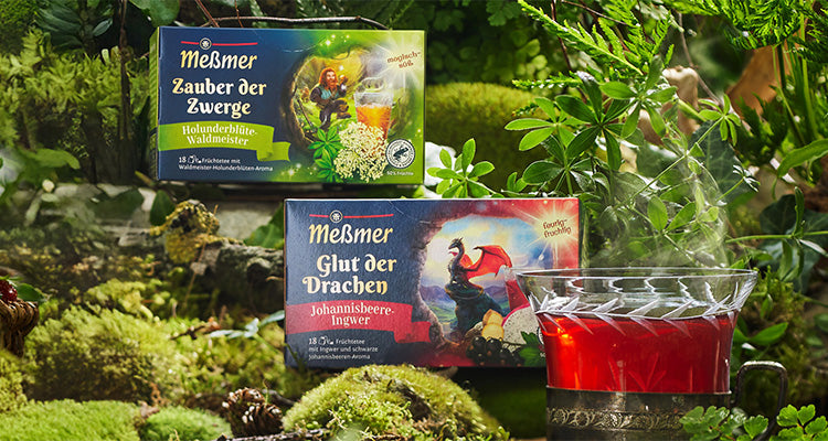 Zwei Schachteln Meßmer-Tee mit fantasievollen Motiven stehen in einer üppigen, grünen Waldlandschaft neben einer Glastasse mit leuchtend rotem Tee. Moos und Blätter umgeben die Szene.