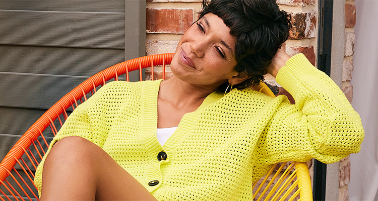 Eine Frau mit kurzen dunklen Haaren in einer leuchtend gelben Strickjacke entspannt sich auf einem orangefarbenen Stuhl, lächelt mit geschlossenen Augen und sitzt im Freien an einer Backsteinmauer.