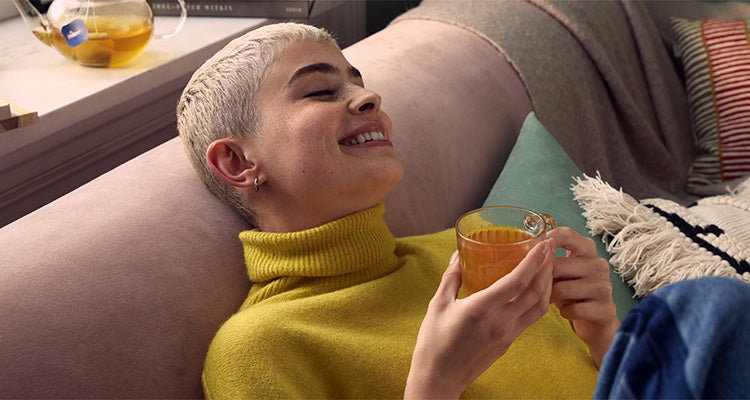 Eine Person mit kurzen blonden Haaren, die einen gelben Rollkragenpullover trägt, lächelt und entspannt sich auf einer Couch, während sie eine Glastasse mit Tee in der Hand hält. Im Hintergrund sind eine Teekanne und Kissen zu sehen.