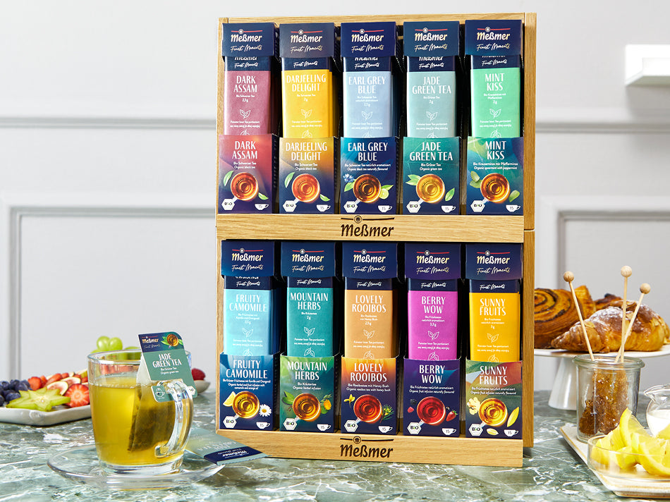 Holzregal mit verschiedenen Meßmer Tee-Sorten in bunten Packungen, darunter Earl Grey, Grüner Tee und Früchtetees, auf Marmoroberfläche mit Tasse, Obst und Gebäck im Hintergrund.