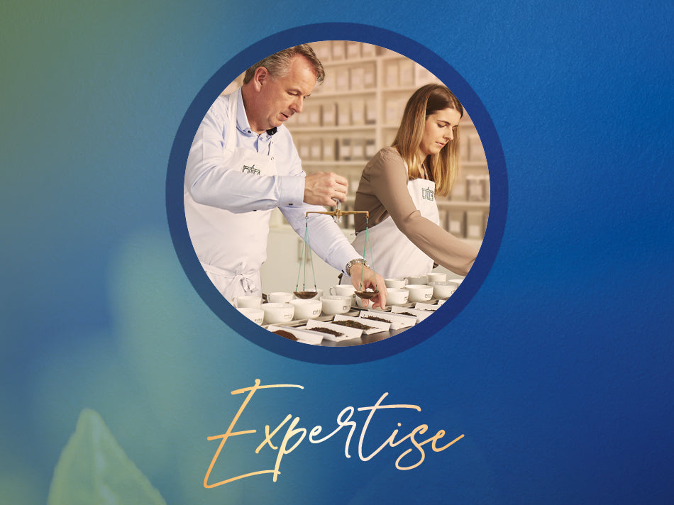 Zwei Personen in weißen Schürzen prüfen sorgfältig Teeproben in kleinen Schalen in einem Labor mit Regalen voller Teepackungen im Hintergrund.