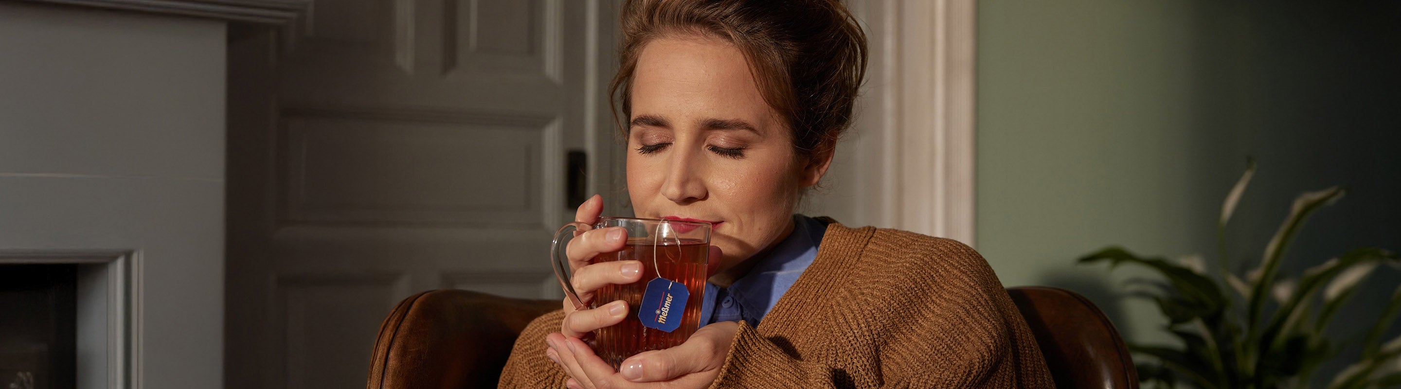 Eine Frau sitzt in einem Haus, die Augen geschlossen und lächelt sanft, während sie mit beiden Händen eine durchsichtige Teetasse hält. Sie trägt einen braunen Pullover, und im Hintergrund sind eine Pflanze und eine Tür zu sehen.