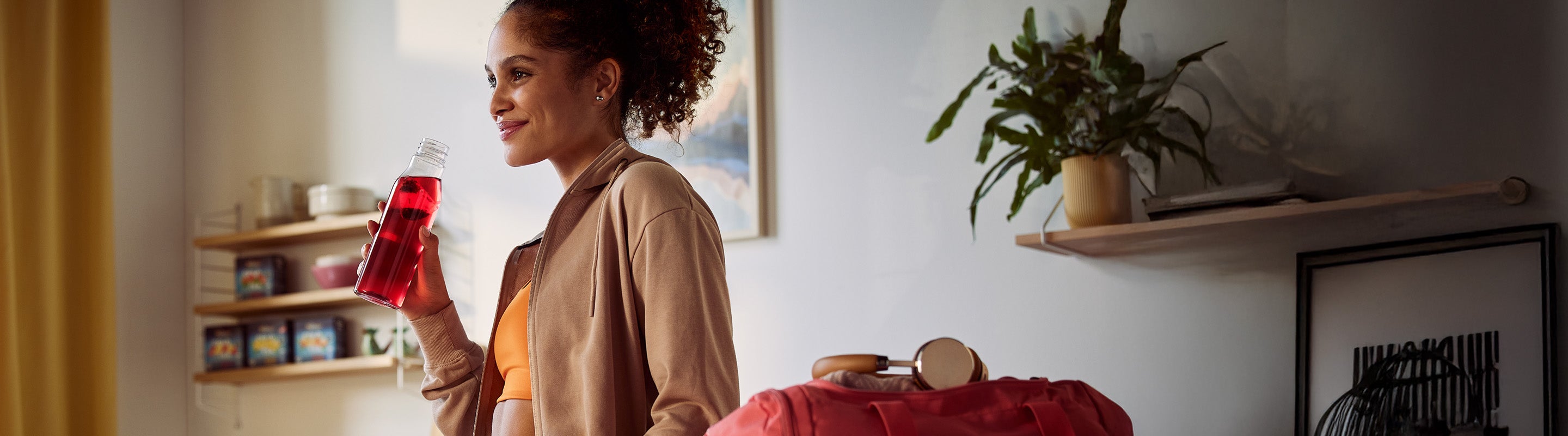 Eine Frau in Sportkleidung steht in einem Raum, hält eine Flasche mit einem roten Getränk in der Hand und lächelt. Eine Sporttasche und eine Pflanze stehen in der Nähe auf einem Regal in dem schwach beleuchteten Raum.