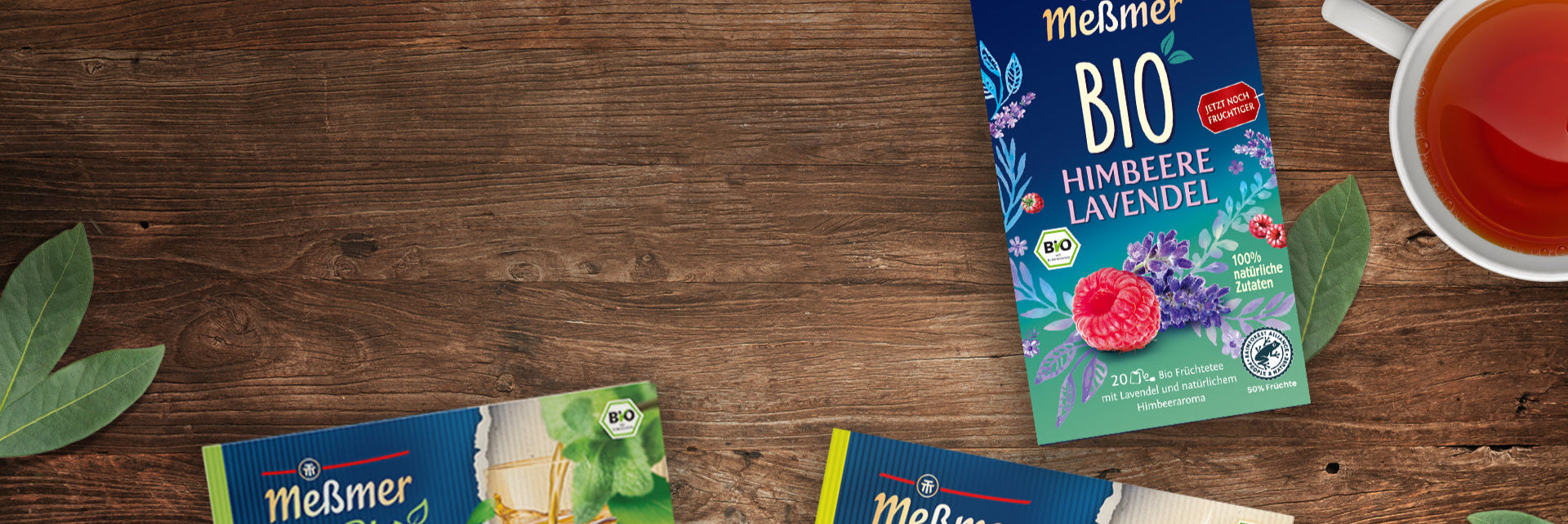 Eine teilweise geöffnete Schachtel Meßmer BIO Himbeere Lavendel-Tee steht auf einem Holztisch neben einer Tasse Tee, Teeblättern und anderen Meßmer Tee-Verpackungen. Das Design zeigt Illustrationen von Himbeeren, Lavendel und Blumen.