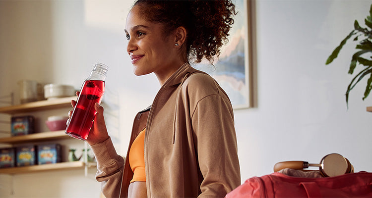 Eine Frau in hellbrauner Jacke und orangefarbenem Oberteil lächelt, während sie eine klare, mit roter Flüssigkeit gefüllte Flasche in der Hand hält und daraus trinkt. Sie steht in einem gemütlichen, sonnendurchfluteten Raum mit Regalen und einer Pflanze im Hintergrund.