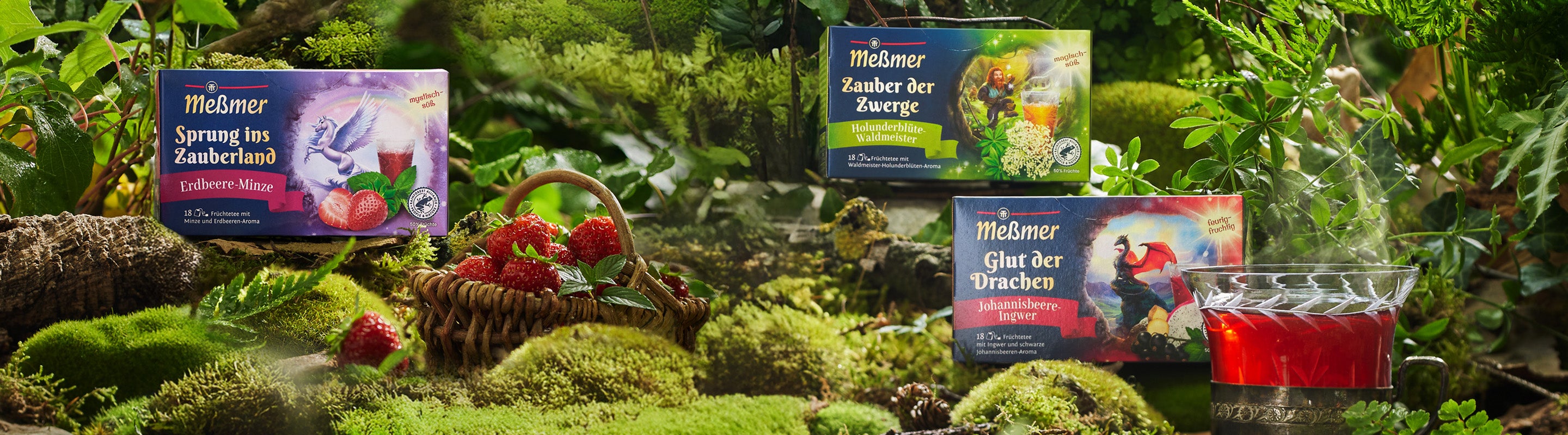 Kisten mit Meßmer-Fantasie-Tee stehen auf Moos mit Erdbeeren, einem Weidenkorb und einer Tasse roten Tees, umgeben von lebhaftem, üppigem Grün in einer magischen Waldumgebung.
