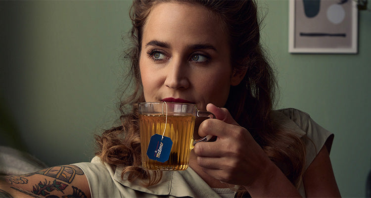 Eine Frau mit gewelltem Haar und Tattoos nippt an einem Tee aus einem klaren Glasbecher mit blauem Teebeutel-Etikett. Sie blickt nachdenklich zur Seite und sitzt drinnen vor einer gedämpften grünen Wand mit gerahmter Kunst.