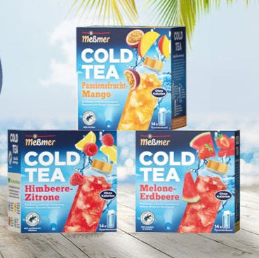 Drei Schachteln Meßmer Cold Tea sind im Freien ausgestellt. Zu den Geschmacksrichtungen gehören Passionsfrucht-Mango, Himbeer-Zitrone und Melone-Erdbeere, jeweils mit Eis und Fruta-Garnierungen. Im Hintergrund sind Palmenblätter und ein blauer Himmel zu sehen.
