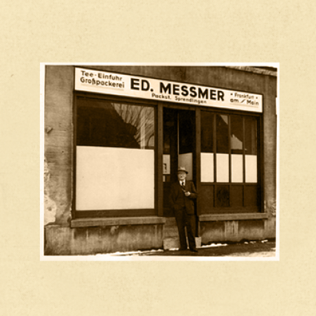 Ein sepiafarbenes Foto eines Mannes mit Hut und Mantel, der vor einem Bäckereifachgeschäft mit der Aufschrift ED. MESSMER auf Deutsch beschriftet ist, mit großen Fenstern und einem Schild über der Tür. Auf dem Boden ist Schnee zu sehen.